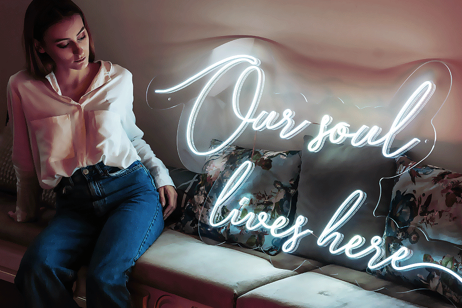 Valkoinen LED-neonteksti “Our soul lives here” sohvan yläpuolella, vieressä istuu nainen